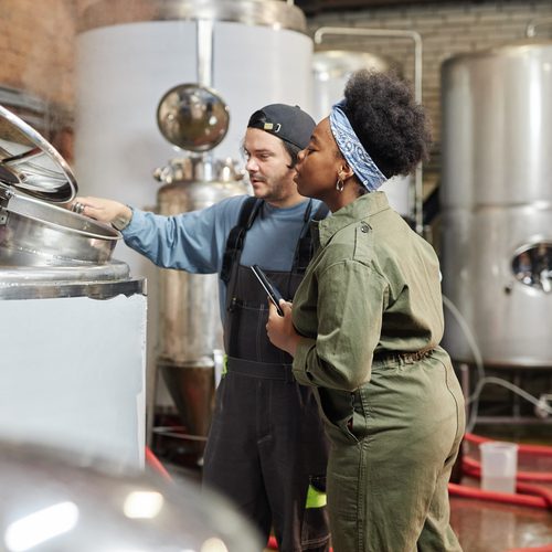 Medium shot of male factory worker opening lid of tank for female African American process technician to observe cider fermentation during quality inspection at plant, copy space