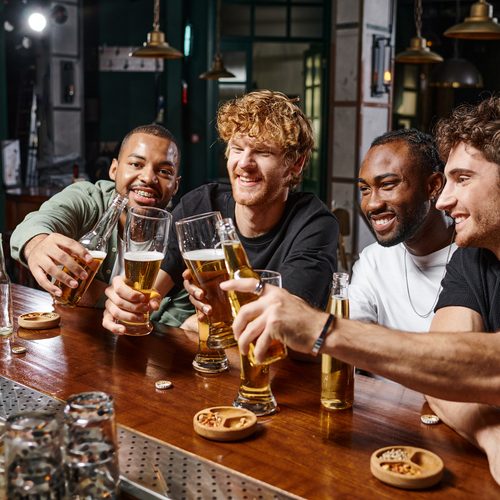 group of four happy multiethnic male friends toasting with glasses of beer at bar counter